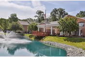 Brick senior living facility building with a covered entrance and deck beside a pond with a fountain and surrounding trees.