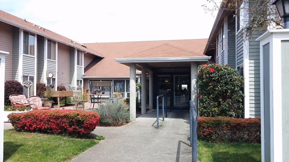 Exterior view of the entrance to Cypress Gardens senior living facility with a covered walkway, surrounded by well-maintained bushes, flowers, and outdoor seating areas.