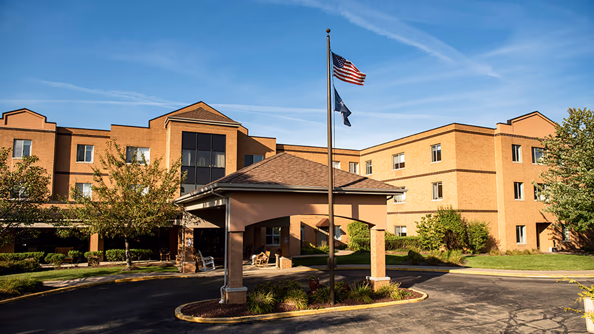 Exterior view of a three-story brick building with a covered entrance and two flagpoles flying the American flag and another flag, surrounded by trees and landscaping under a clear blue sky.