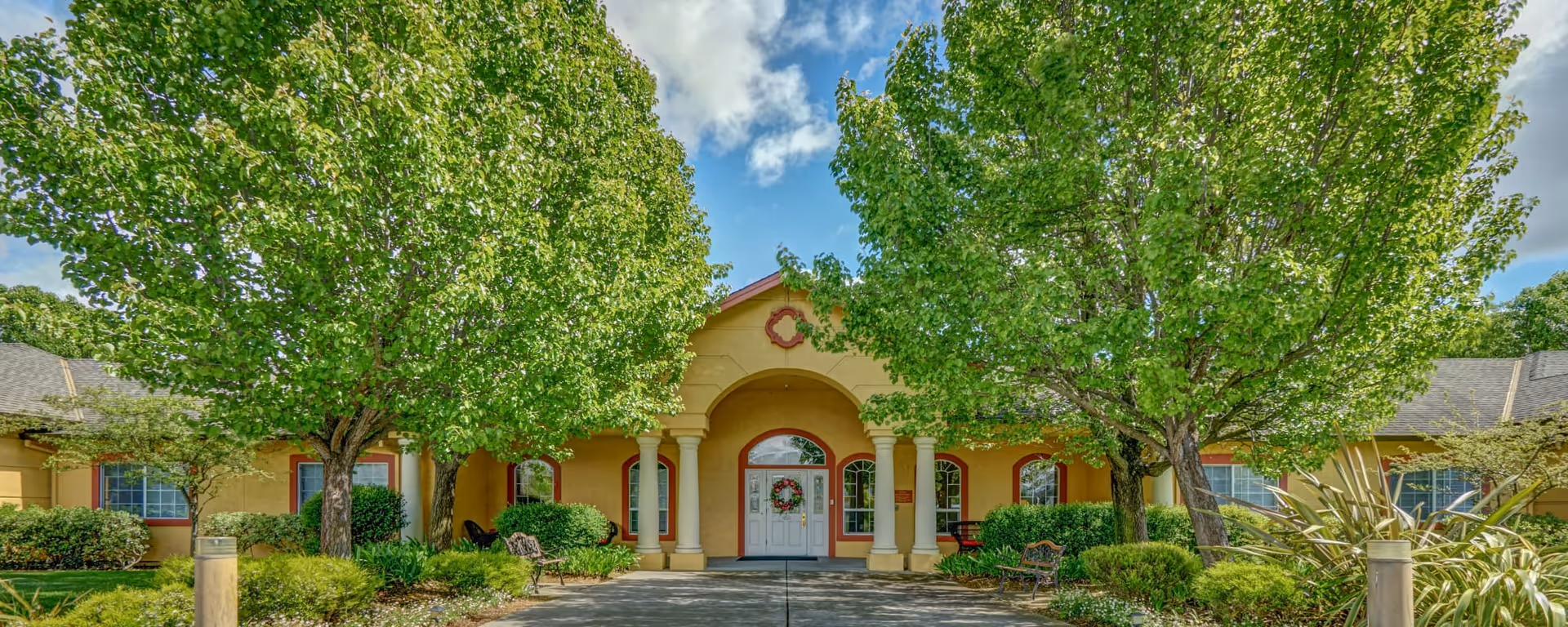 Front exterior view of MuirWoods Memory Care facility with a yellow building, white double doors decorated with a wreath, surrounded by green trees and shrubs under a partly cloudy blue sky.