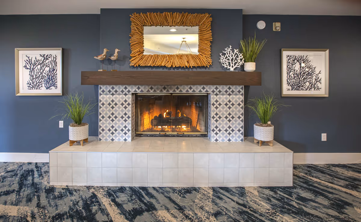 Stylish indoor fireplace with a driftwood mirror above, patterned tile surround, potted plants, and framed wall art on a dark blue wall.