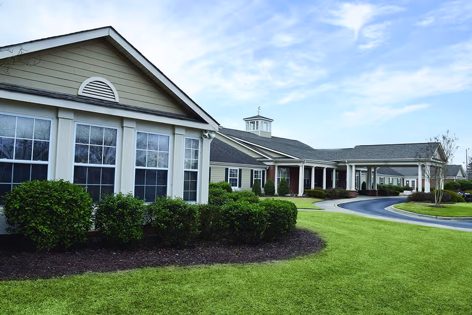 Exterior view of Spring Arbor of Greenville senior living facility showing a single-story building with beige siding, multiple windows, a covered entrance with white columns, neatly trimmed bushes, and a well-maintained lawn under a partly cloudy sky.