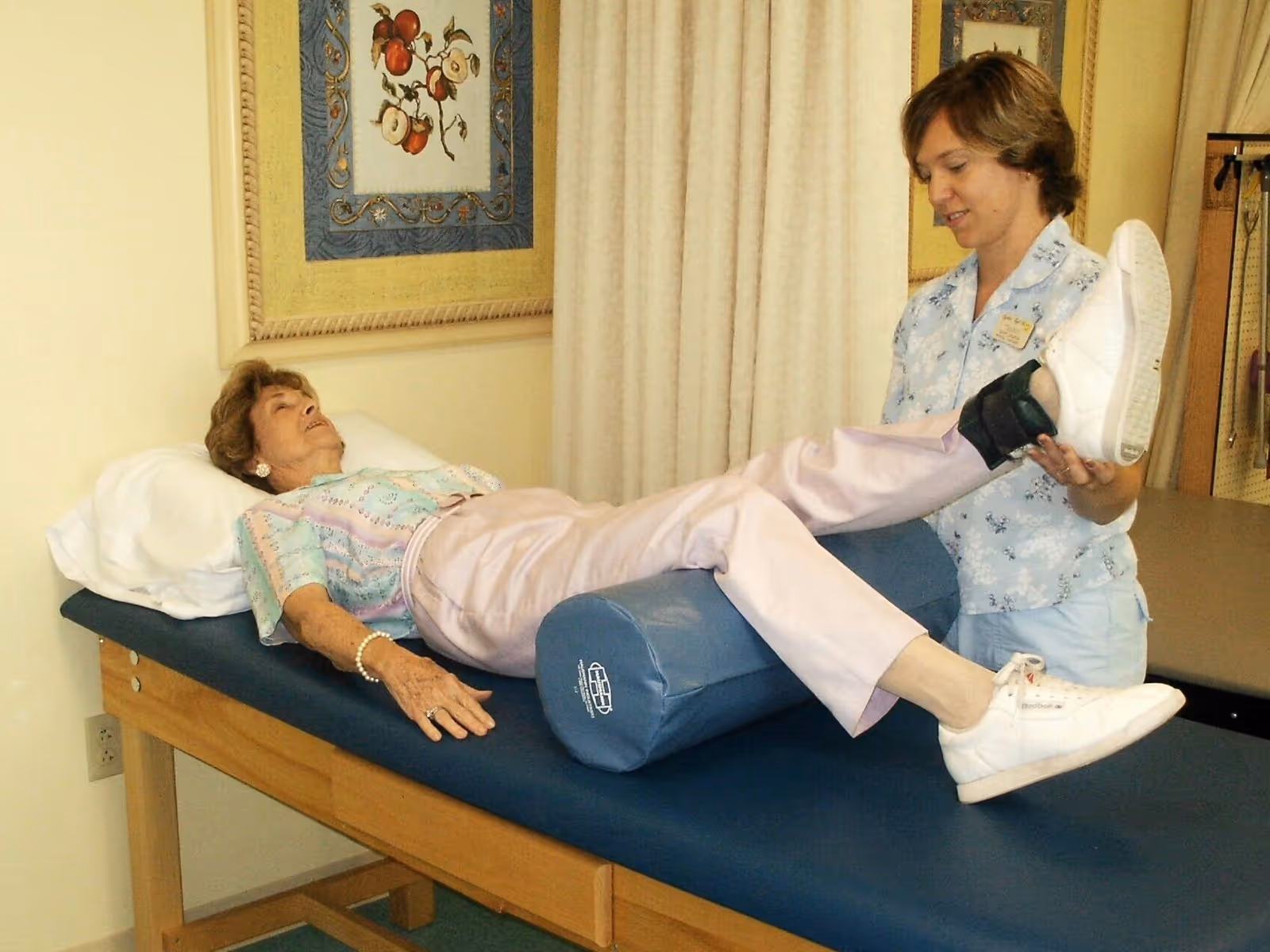 An elderly woman lies on a padded therapy table with a pillow under her knees while a healthcare worker assists by holding and lifting one of her legs during a physical therapy session in a rehabilitation center.