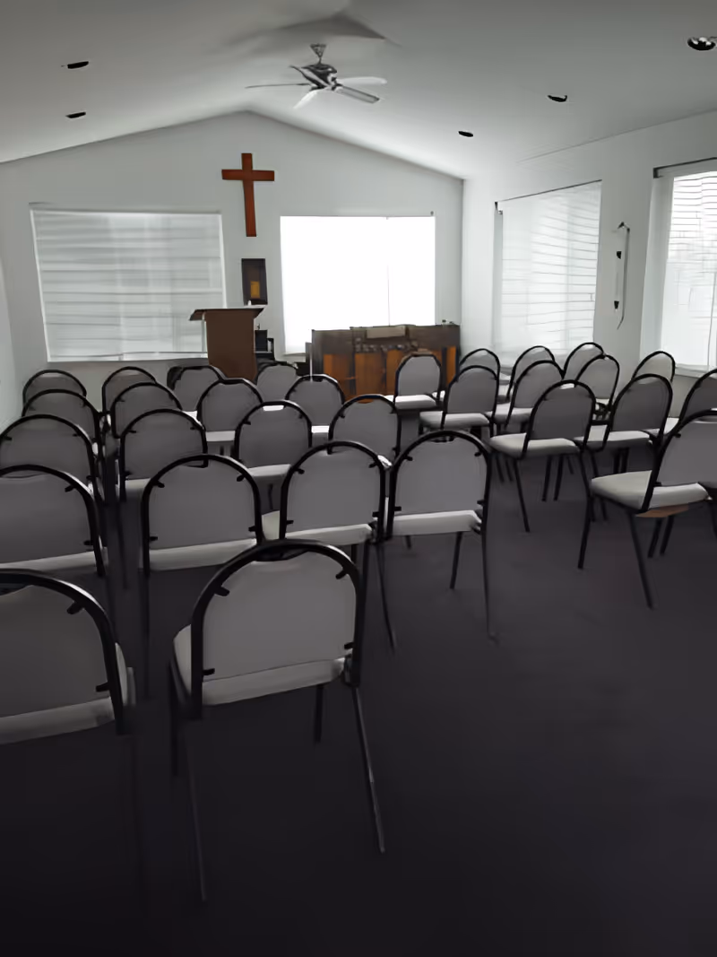 Small chapel-style meeting room with rows of chairs facing a podium, piano, and a wooden cross on the front wall.
