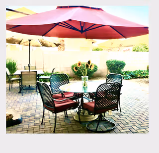 Outdoor patio area with a round glass table surrounded by four metal chairs with red cushions. A large red umbrella provides shade over the table. In the background, there is another table with chairs and a smaller umbrella, along with a brick wall and some greenery including bushes and sunflowers.