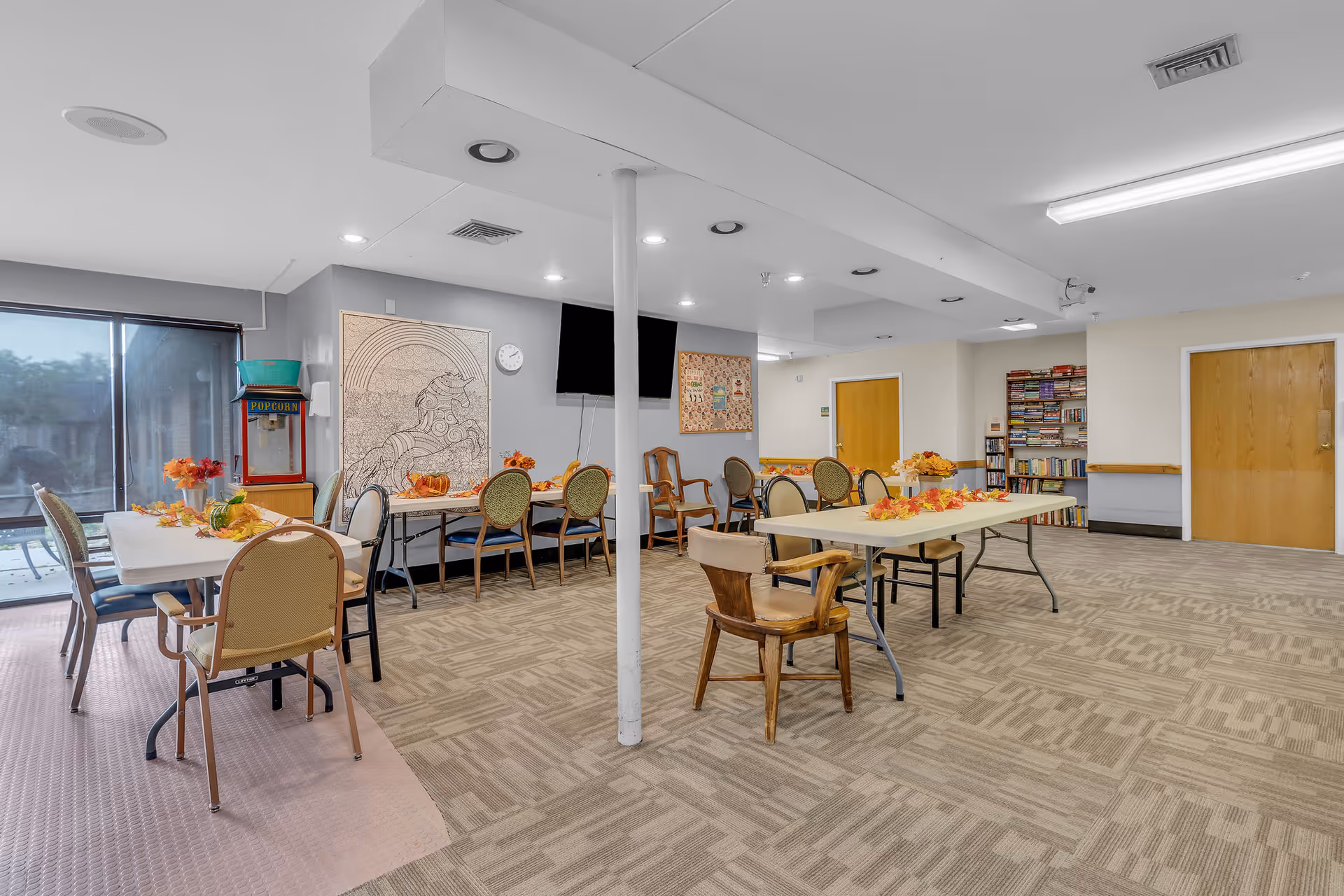 A spacious common area in a senior living facility with several tables and chairs arranged for group activities or dining. The room features a popcorn machine, a large window with a view outside, a wall-mounted TV, a bookshelf filled with books, and autumn-themed decorations on the tables.