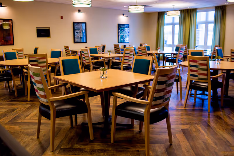 A bright dining room with multiple wooden tables and chairs featuring colorful patterned backs and teal cushions. The room has large windows allowing natural light to fill the space, wooden flooring, and framed artwork on the walls.