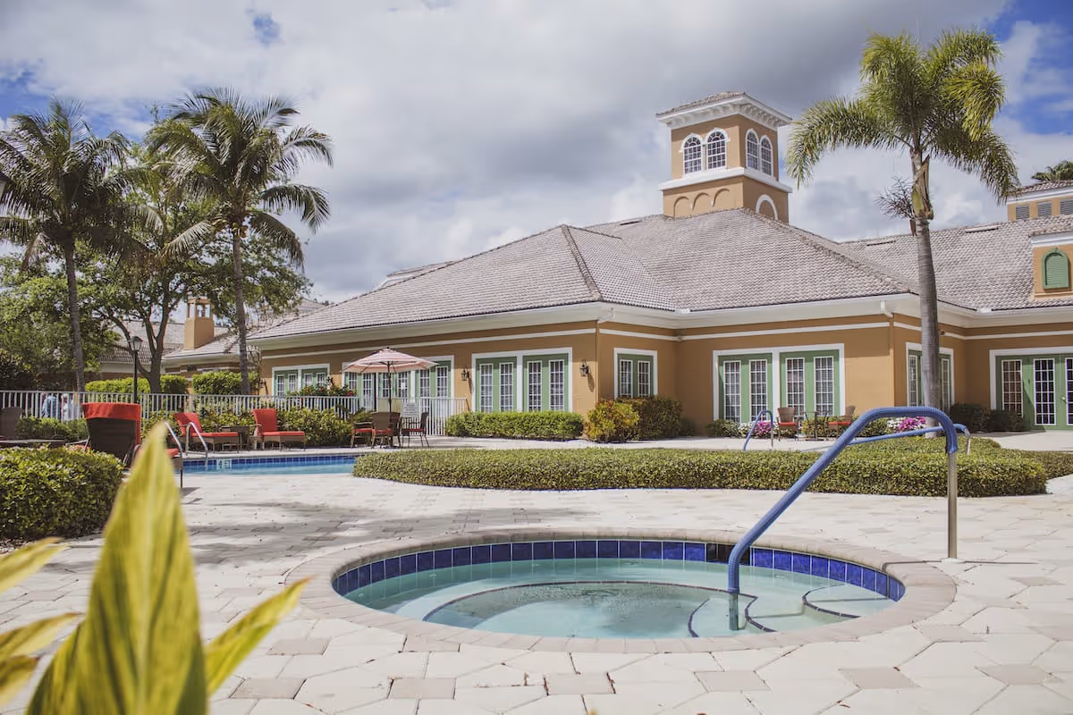 Outdoor view of a senior living facility with a hot tub in the foreground, a swimming pool with red lounge chairs and umbrellas, palm trees, and a large building with multiple windows and a tower under a partly cloudy sky.