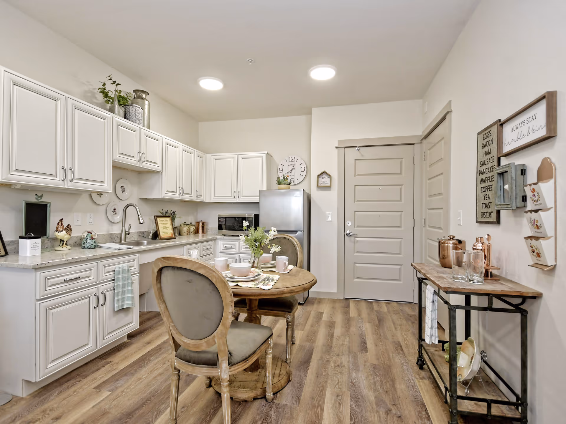 A bright kitchen area in a senior living facility featuring white cabinets, a stainless steel refrigerator, a round wooden dining table set with dishes and cups, wooden chairs with cushioned seats, and decorative items on the walls and countertops. The floor is wood-style laminate, and the room has a clean, cozy atmosphere.