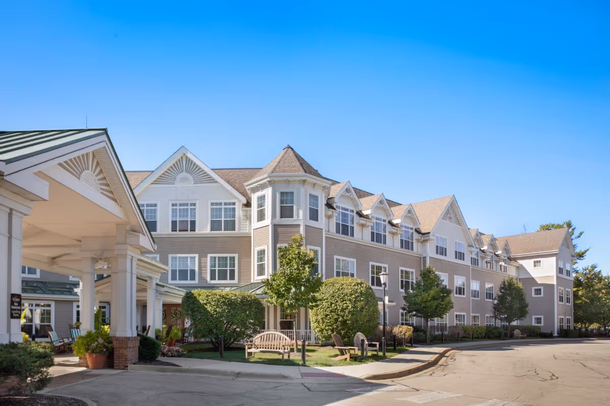 Three-story senior living building with a covered entrance, benches and landscaping under a clear blue sky.