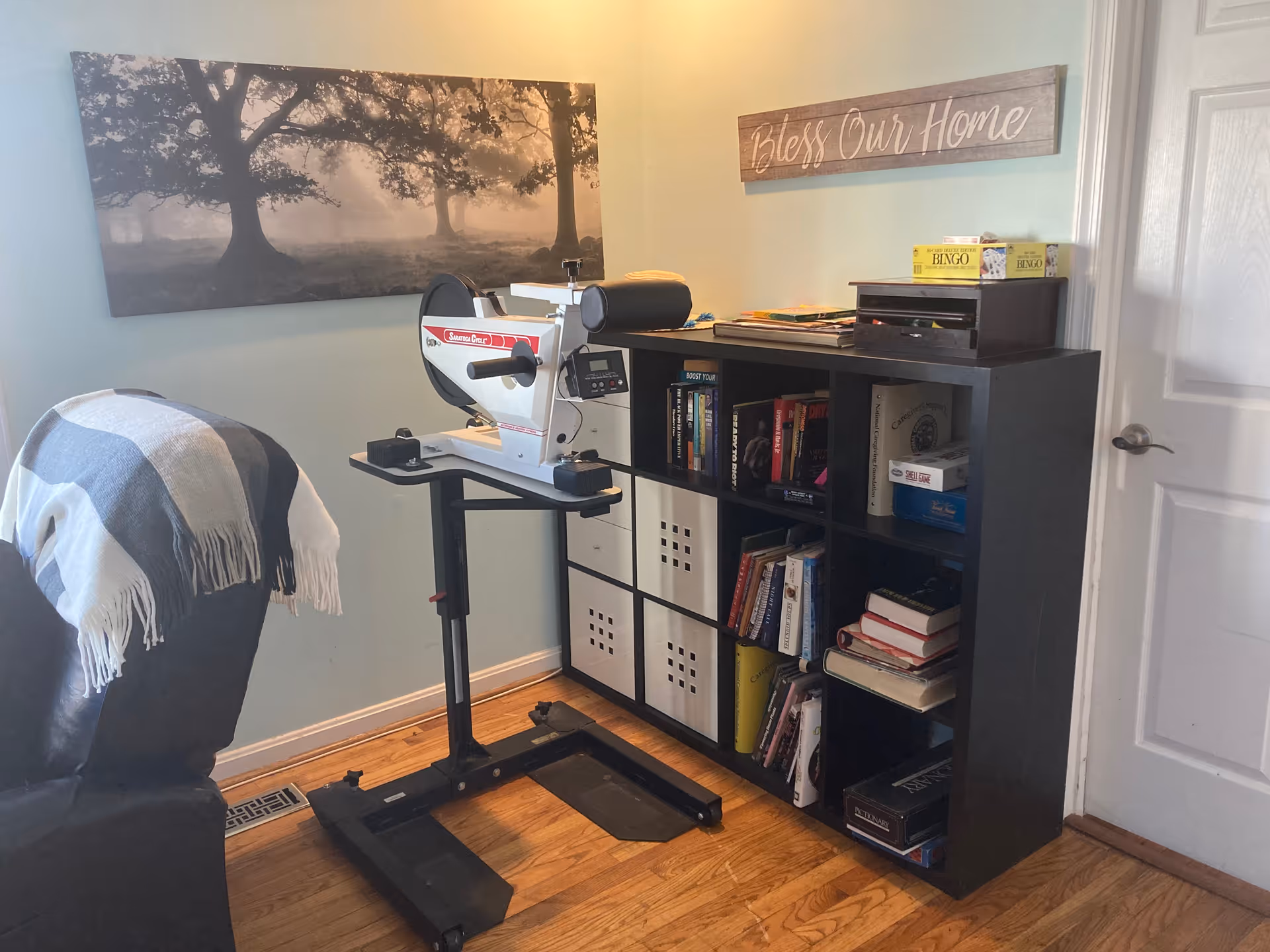Corner of a living room showing a bookshelf with books and boxes, a medical device on a rolling stand, a recliner draped with a blanket, and a 'Bless Our Home' sign on the wall.