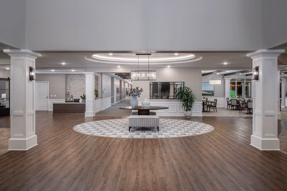 Spacious and modern interior lobby area with wood flooring, white columns, a round patterned rug, a central table with decorative vases and plants, and a seating bench. The area is well-lit with ceiling lights and features a hallway and dining area in the background.