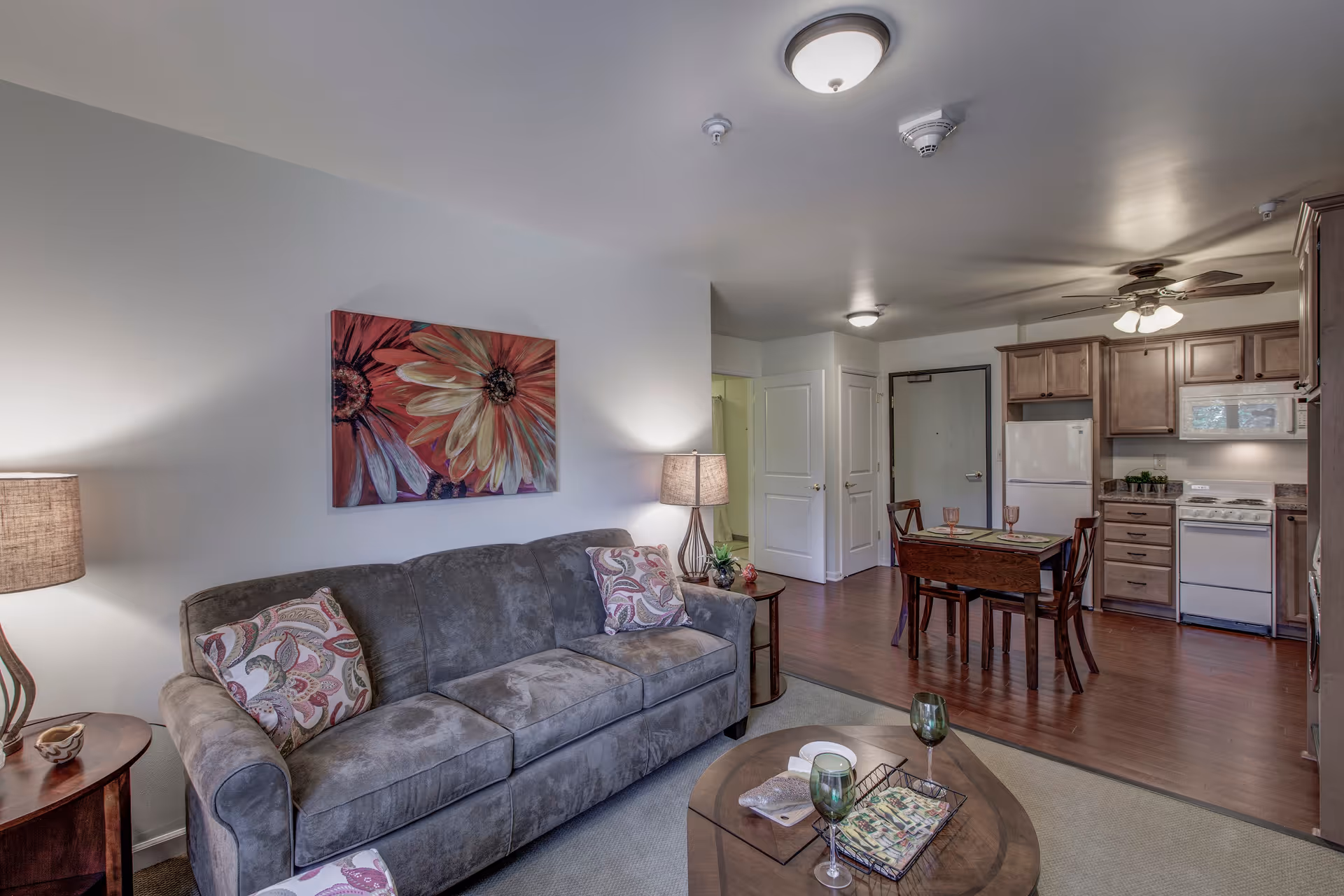 A cozy living room and kitchen area in an apartment at Elizabeth Gardens. The living room features a gray sofa with patterned pillows, a round wooden coffee table with two green wine glasses and a decorative tray, and two side tables with lamps. A colorful floral painting hangs on the wall above the sofa. The kitchen has wooden cabinets, a white refrigerator, stove, and microwave, and a small dining table with four chairs set with placemats and glasses. The floor is a combination of carpet and wood, and the ceiling has a fan and light fixtures.