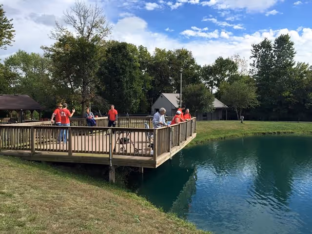 Several people standing and leaning on the wooden railing of a deck overlooking a pond in a grassy outdoor area with trees and a small building in the background under a partly cloudy sky.