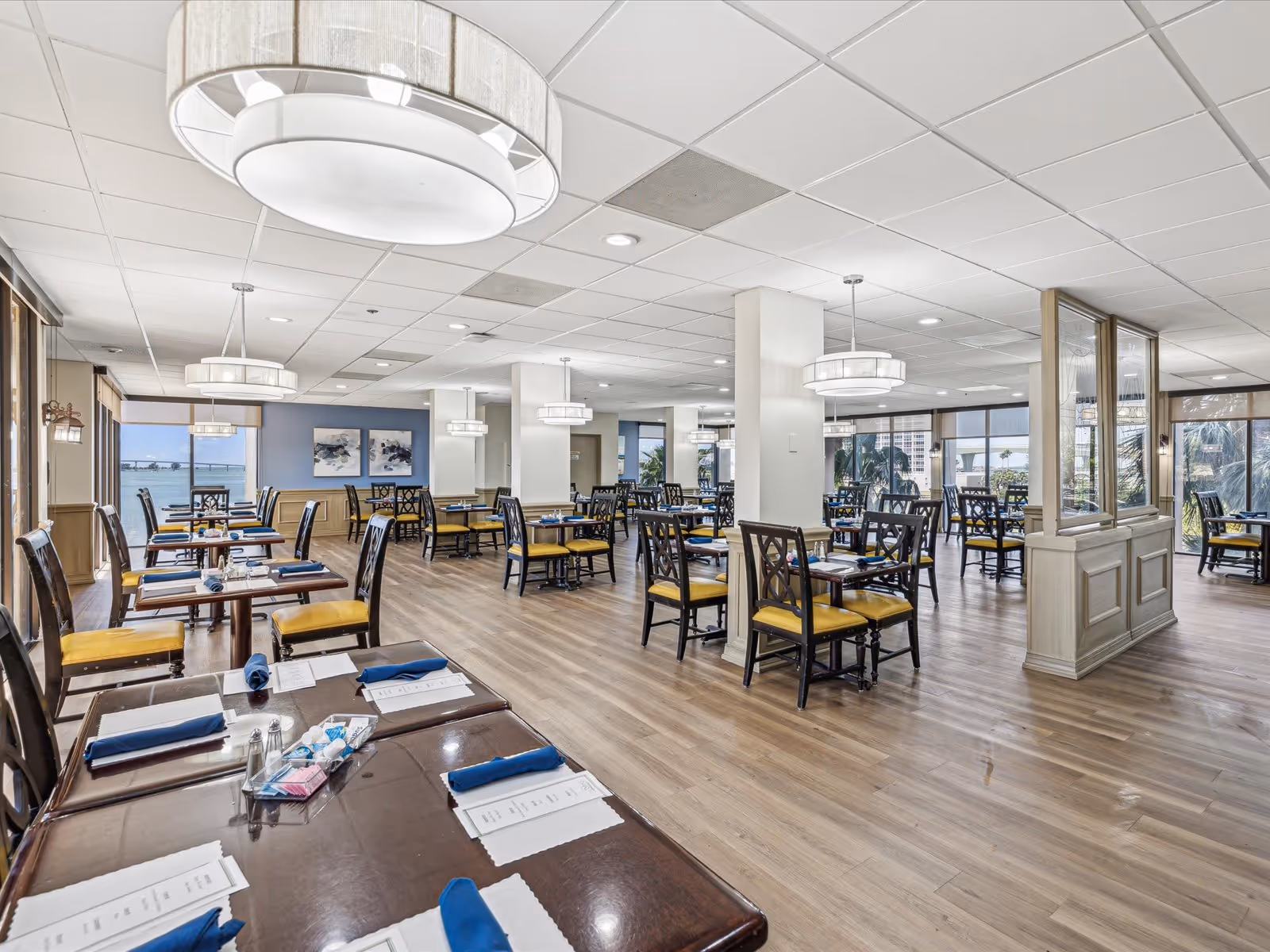 Spacious dining room with multiple wooden tables and chairs featuring yellow cushions, set with blue napkins and menus. Large windows provide a view of palm trees and water outside. Modern ceiling lights hang from a white tiled ceiling, and the floor is covered with light wood laminate.