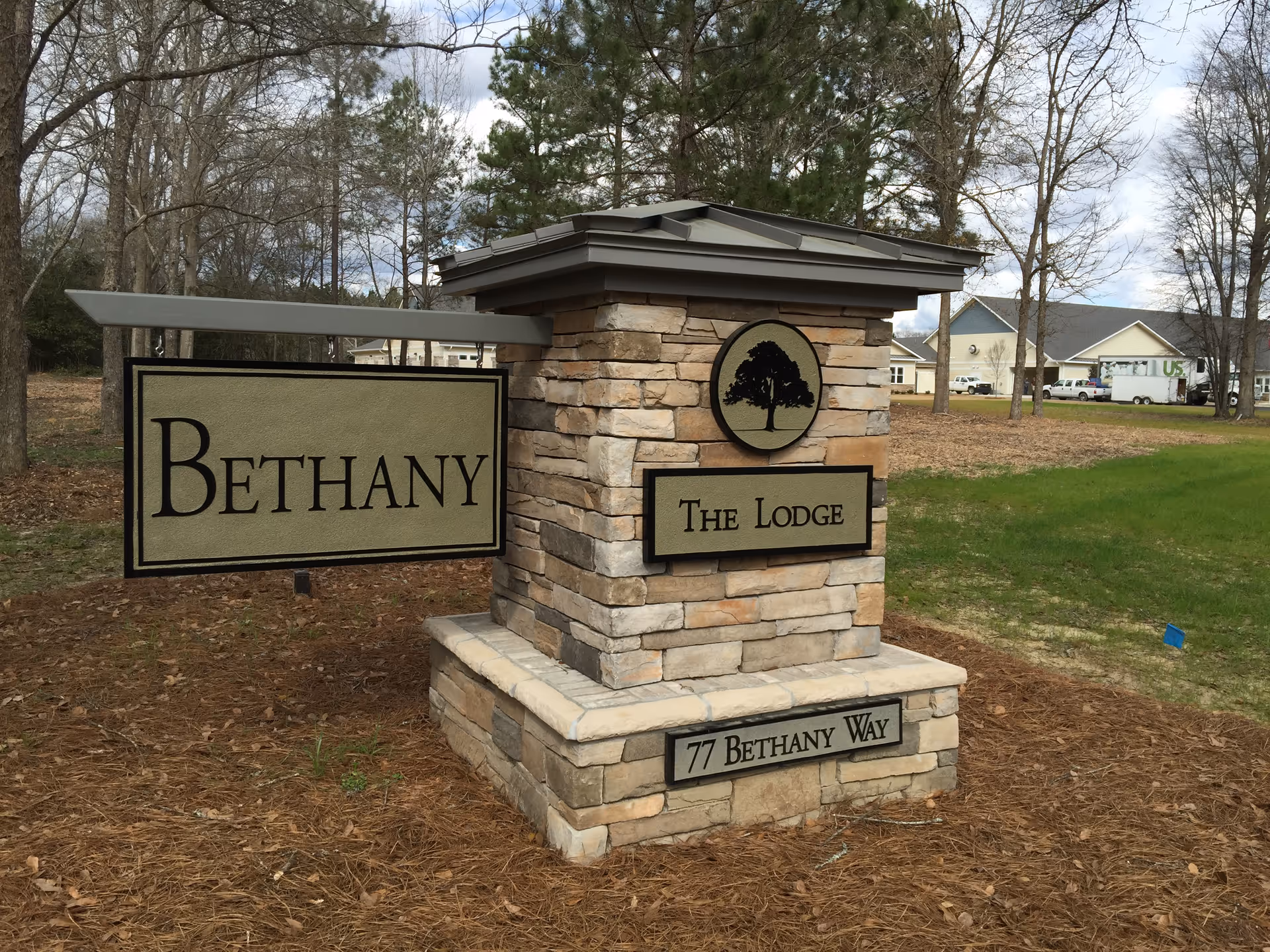 Stone monument sign with a tree logo and text reading 'The Lodge' and 'Bethany' on a hanging sign, located at 77 Bethany Way, surrounded by grass and trees with buildings in the background.