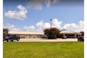 Single-story facility with a covered entrance and parking lot, set behind a grassy lawn under a partly cloudy sky.