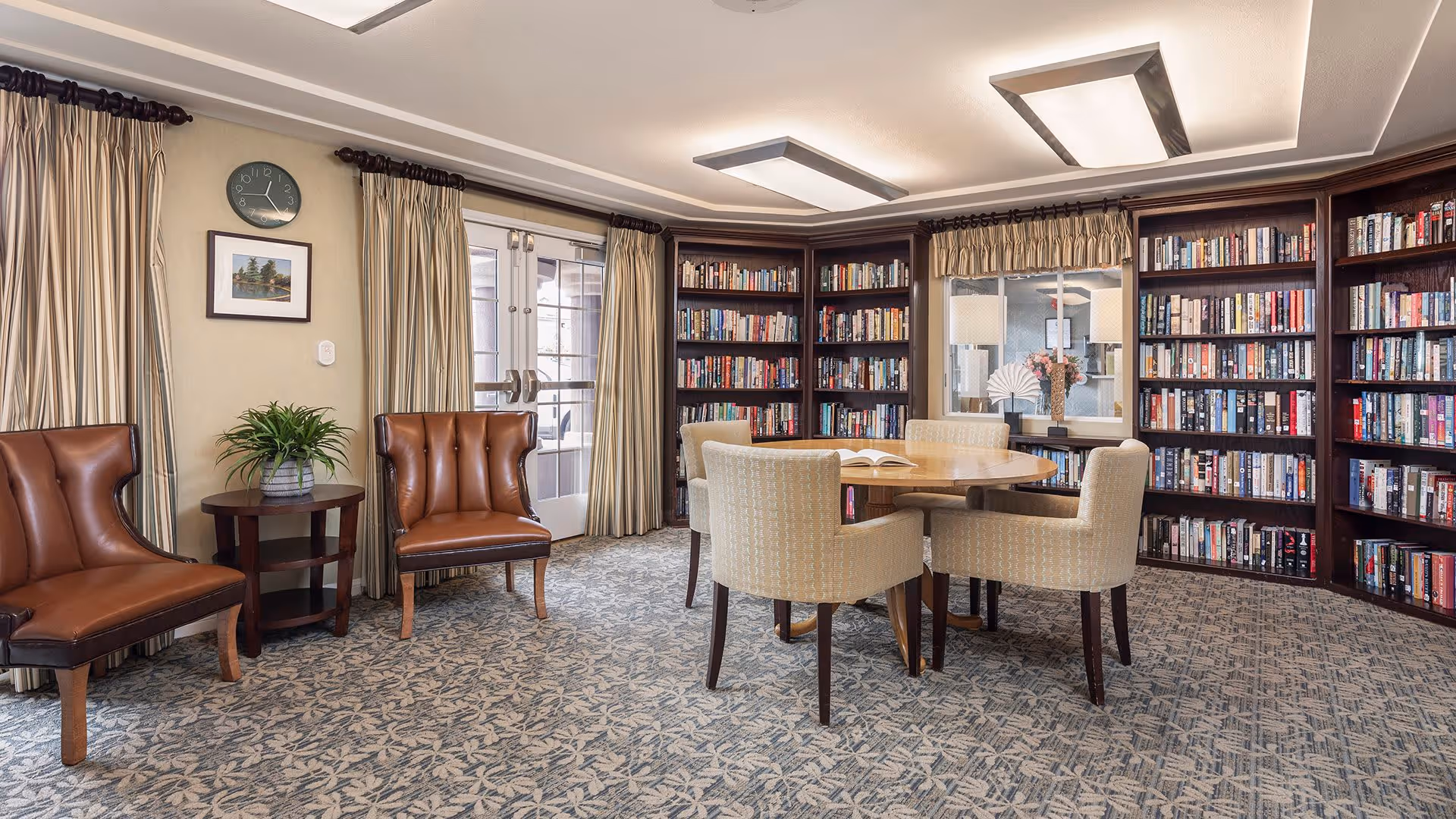 A cozy library room with built-in dark wood bookshelves filled with books along the walls. In the center, there is a round wooden table surrounded by four beige upholstered chairs. Two brown leather chairs flank a small round wooden side table with a potted plant. The room has patterned carpet, beige walls, and large windows with beige curtains. A clock and framed picture hang on the wall above the chairs.