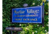 Blue Mayfair Village sign reading Front Entrance, surrounded by lush greenery and shrubs.