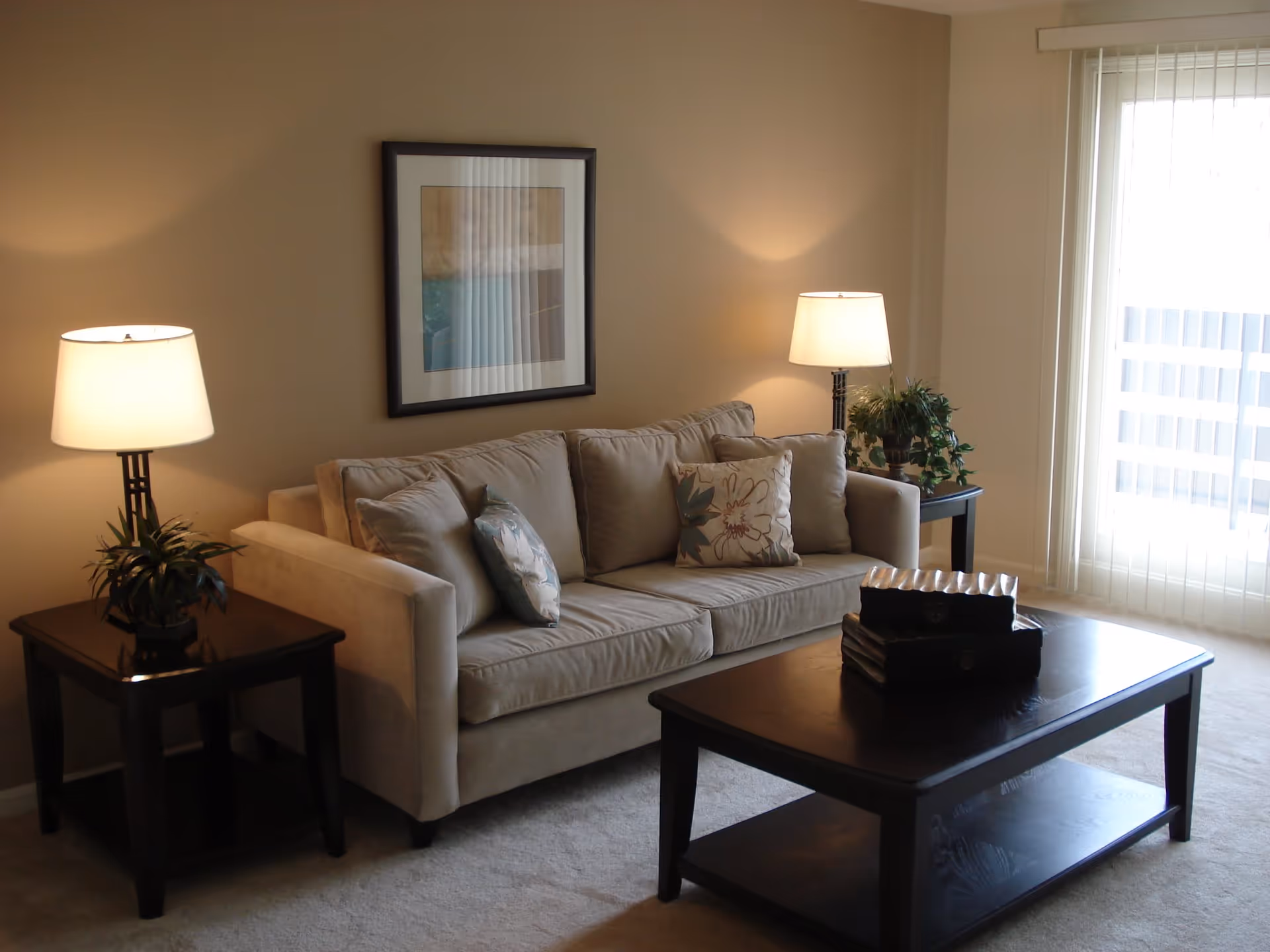 A cozy living room with a beige sofa adorned with three decorative pillows. There are two dark wooden side tables on either side of the sofa, each with a lamp and a small plant. A dark wooden coffee table is in front of the sofa with decorative items on top. A framed abstract artwork hangs on the wall above the sofa. Vertical blinds cover a large window or sliding door, allowing natural light to enter the room.