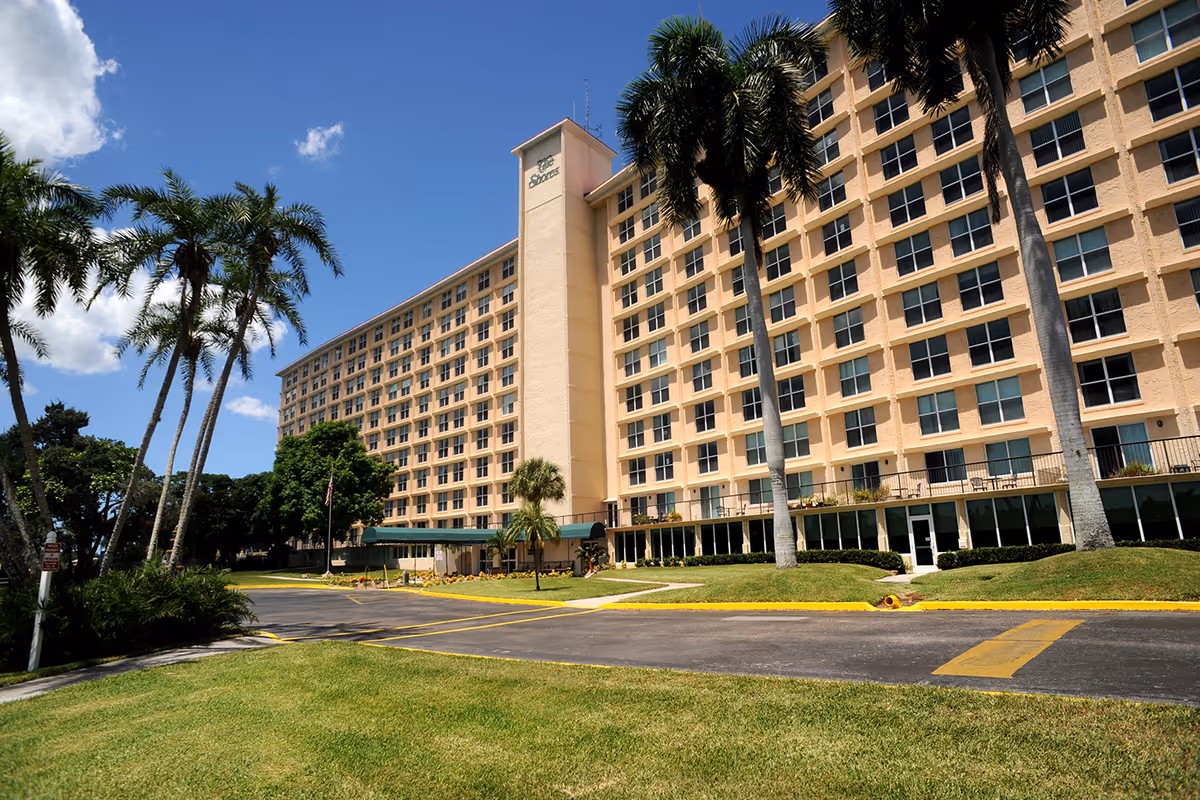 Exterior view of a large multi-story senior living facility building with numerous windows, surrounded by palm trees and green lawns under a clear blue sky.