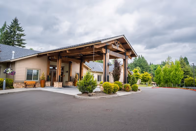 Exterior view of Sunnyside Meadows Memory Care facility entrance with a large wooden covered driveway, surrounded by greenery and trees under a cloudy sky.