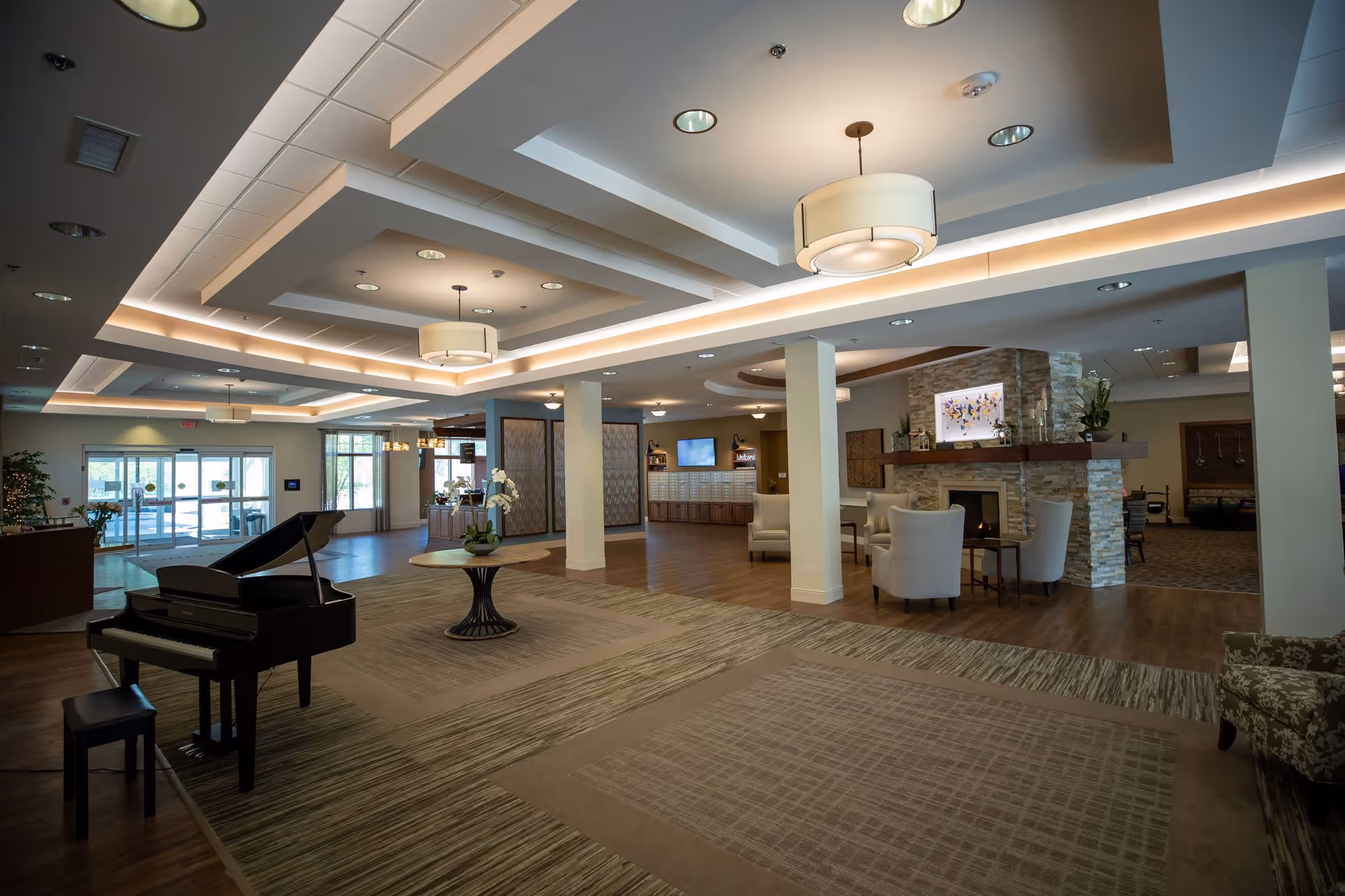 Spacious and well-lit senior living facility common area with a grand piano on a carpeted section, modern ceiling lights, a stone fireplace with a mounted TV above it, comfortable armchairs arranged around the fireplace, and large windows near the entrance allowing natural light.
