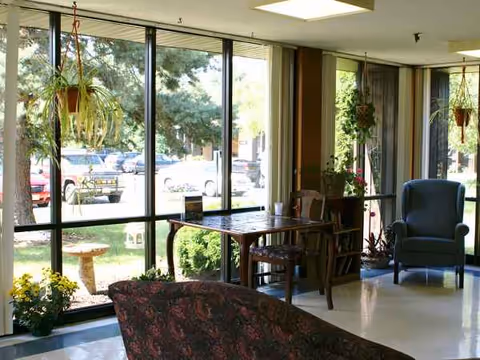 Sunlit senior living lounge with large windows, plants, and seating.