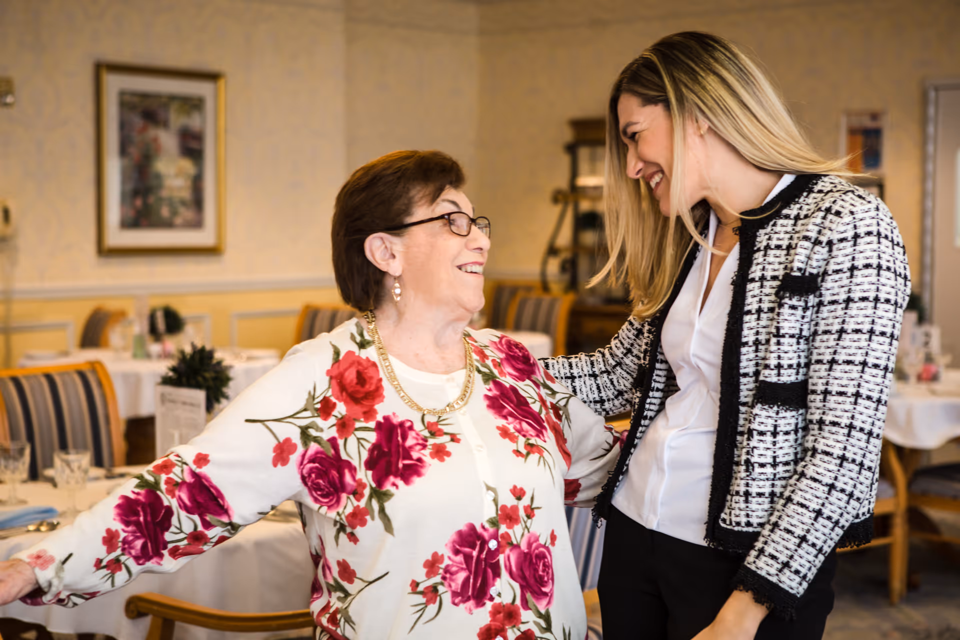 An elderly woman wearing glasses and a floral sweater smiling and interacting warmly with a younger woman in a checkered jacket inside a dining room with tables and chairs in the background.