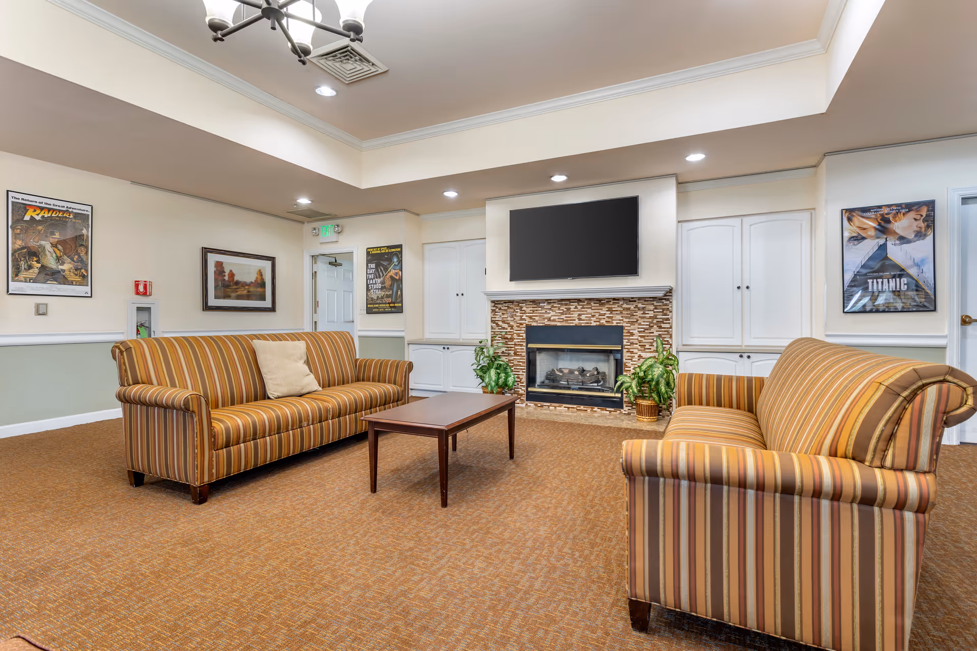 A cozy living room area with two striped sofas facing each other across a wooden coffee table. The room features a fireplace with a mosaic tile surround and a flat-screen TV mounted above it. There are framed movie posters on the walls, including Raiders of the Lost Ark and Titanic, along with a framed landscape painting. The room has recessed lighting and a chandelier, with a carpeted floor and light-colored walls.