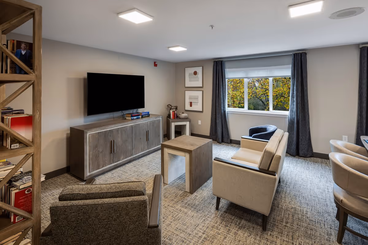 A cozy senior living community lounge with a wall-mounted flat-screen TV above a wooden cabinet, a small wooden coffee table, a beige loveseat, a dark armchair, and a window with dark curtains showing trees outside. There is a bookshelf on the left side filled with books and a small side table with decorative items in the corner.