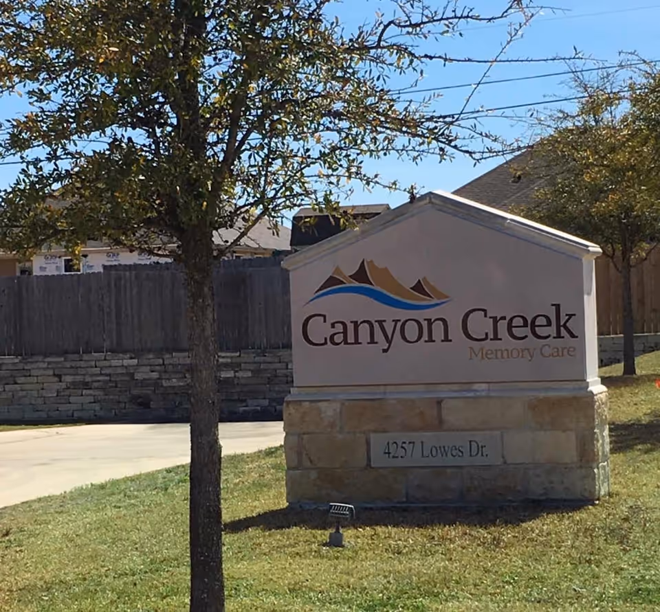 Outdoor view of a stone and concrete sign for Canyon Creek Memory Care located on a grassy area with a tree nearby and a driveway in the background.