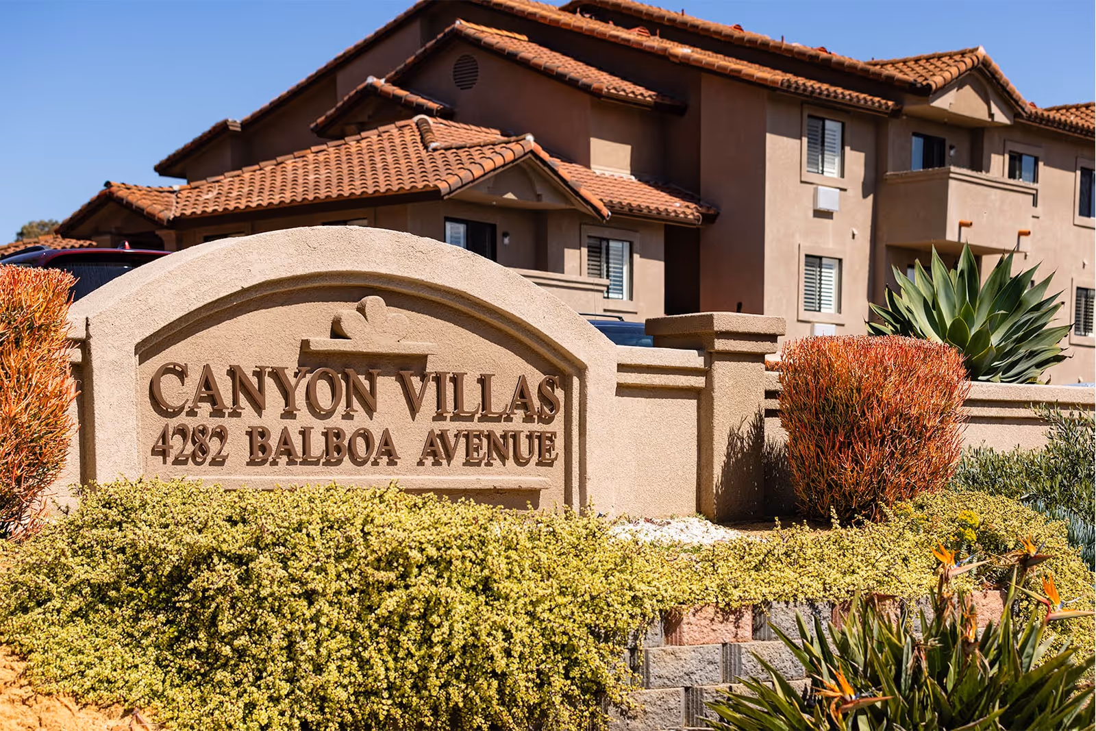 Stone entrance sign reading 'Canyon Villas 4282 Balboa Avenue' with landscaped shrubs and a stucco building with tile roofs behind it.