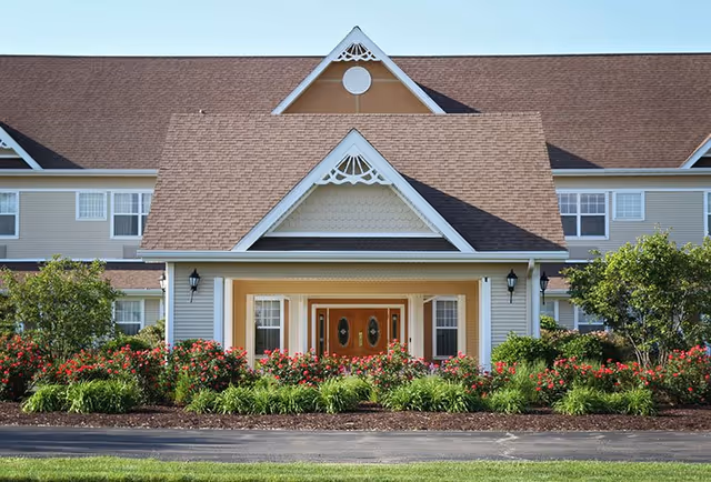 Front exterior view of a senior living facility building with a peaked roof, beige siding, white trim, and a wooden double door entrance. The entrance is surrounded by well-maintained landscaping with green bushes and red flowers.