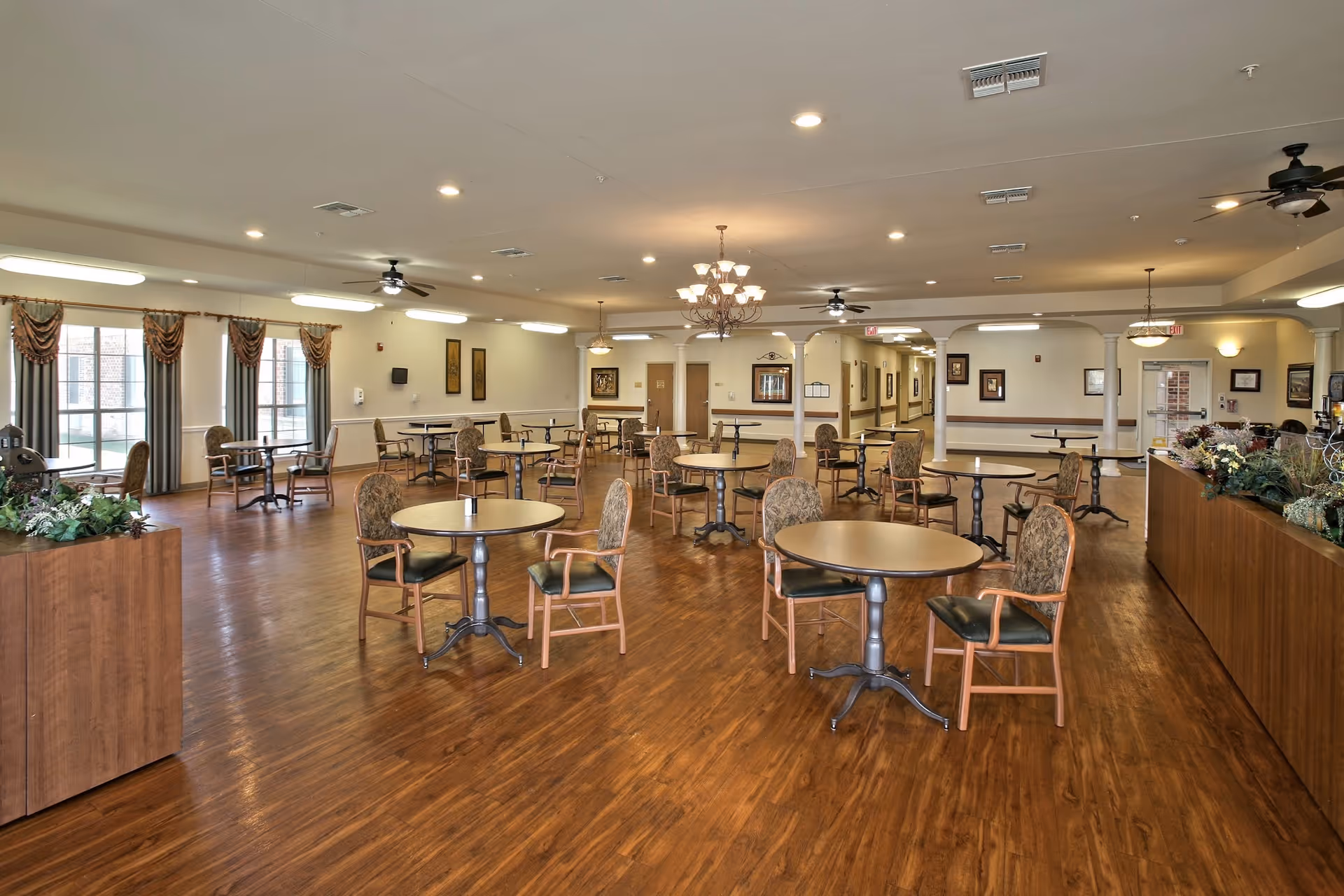 Spacious dining room in a senior living facility with multiple round tables and chairs arranged neatly on a wooden floor. The room is well-lit with ceiling lights and chandeliers, and large windows with curtains allow natural light to enter. There are decorative plants and framed artwork on the walls, and a counter area on the right side.