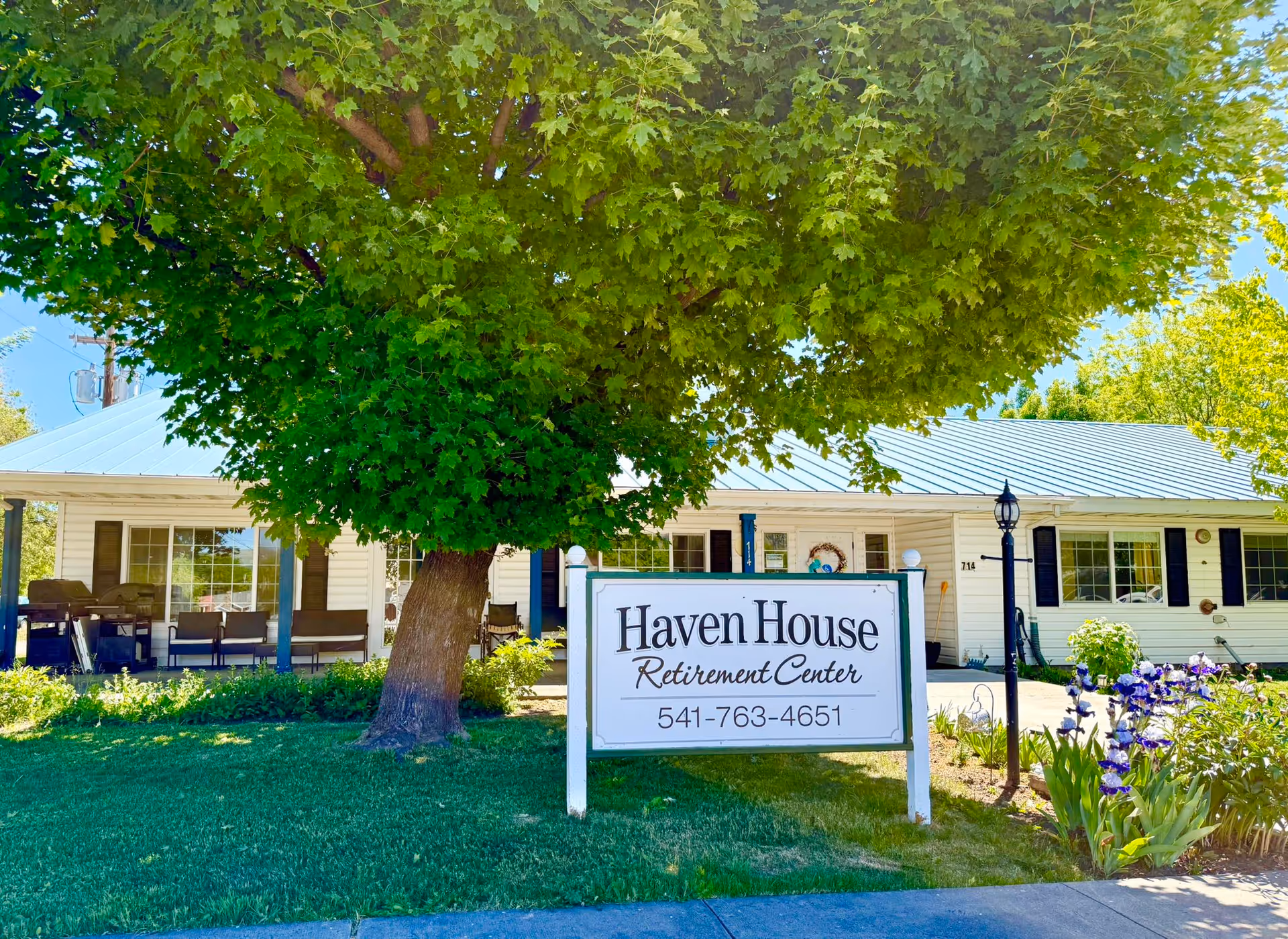 Front exterior view of Haven House Retirement Center with a large leafy tree in front, a white sign displaying the facility name and phone number, and a single-story building with white siding and black shutters in the background.