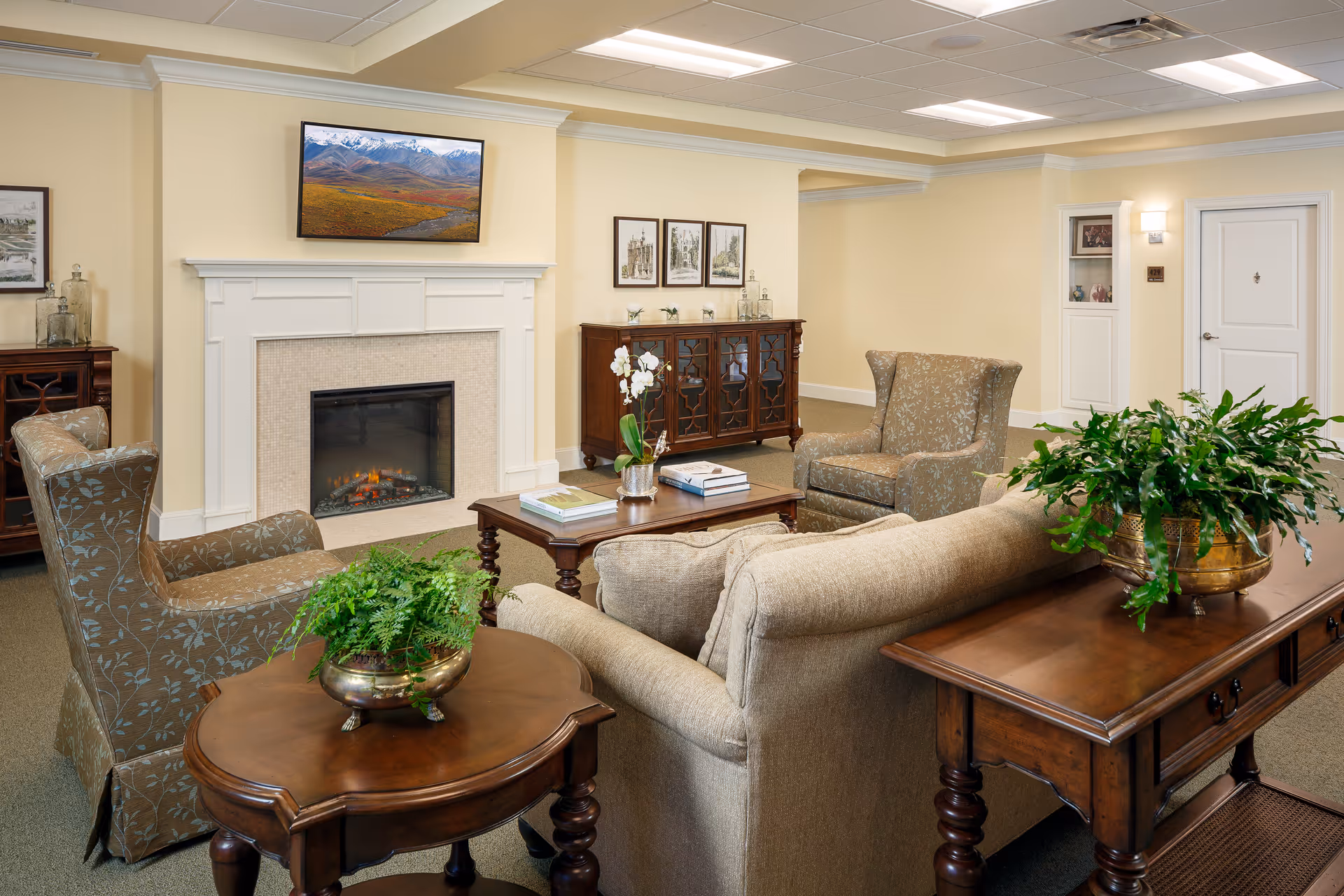 A cozy living room area in an assisted living facility featuring a beige sofa, two patterned armchairs, a wooden coffee table with books and a small plant, a fireplace with a mounted TV above it displaying a mountain landscape, and additional wooden furniture with decorative items and plants. The walls are painted light yellow and the room is well-lit with ceiling lights.