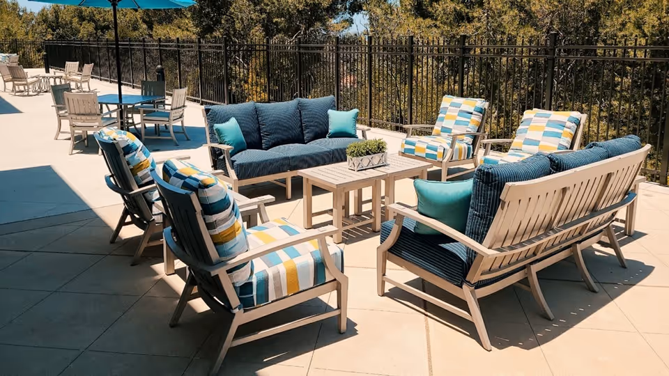 Outdoor patio area with cushioned seating including two sofas and four armchairs arranged around a rectangular coffee table. The chairs have colorful striped cushions in blue, yellow, and white, while the sofas have solid blue cushions with teal accent pillows. There are additional tables and chairs in the background under large blue umbrellas, with a black metal fence and trees beyond.