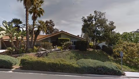 Single-story house with a tiled roof on a landscaped slope, surrounded by palm trees and shrubs along the street.