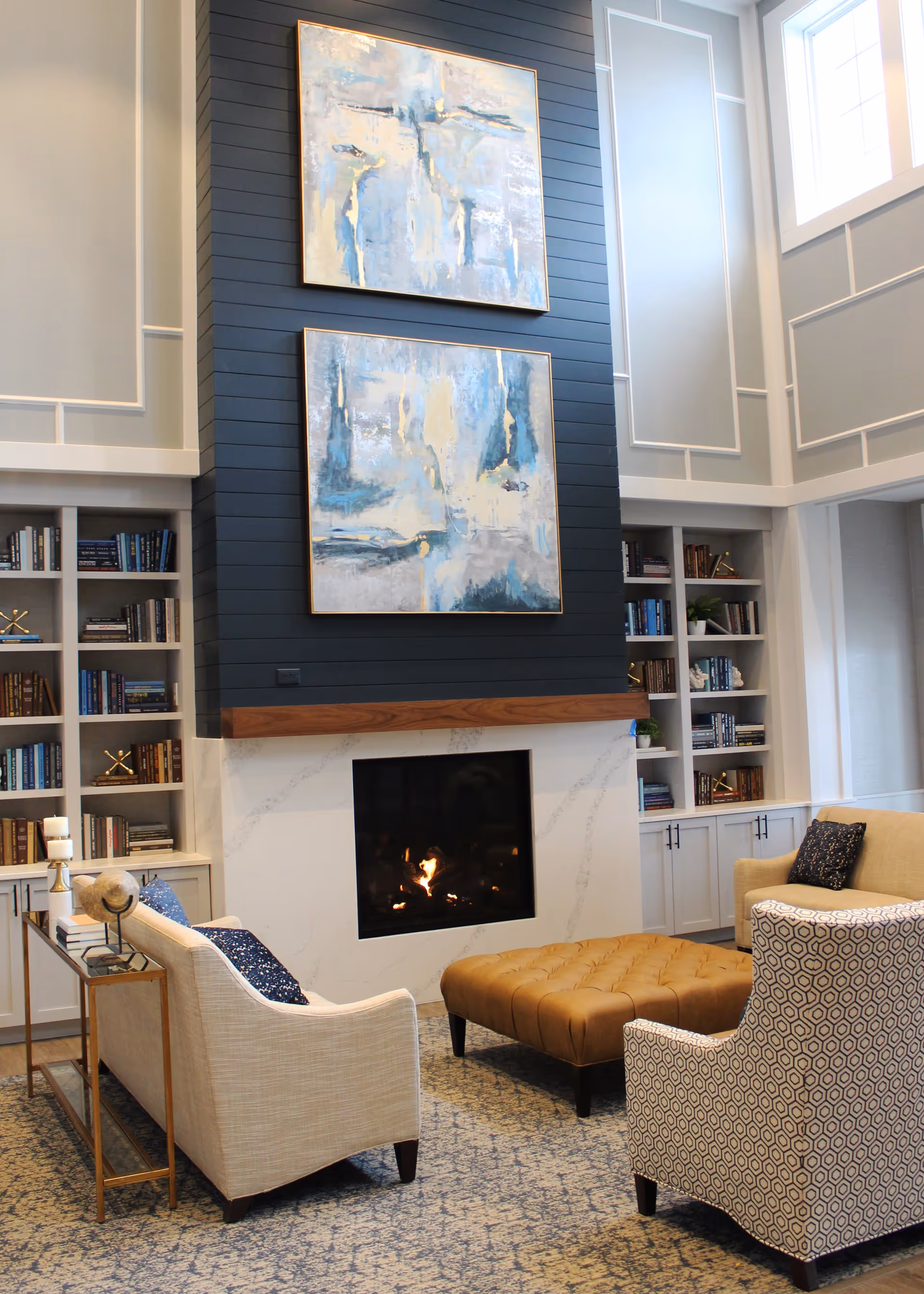 A cozy living room area with a modern fireplace set in a white marble surround and a dark blue shiplap accent wall. Above the fireplace are two large abstract paintings in shades of blue, white, and gold. The room features built-in white bookshelves filled with books and decorative items on either side of the fireplace. Comfortable seating includes a beige sofa with blue patterned pillows, a patterned armchair, and a large tufted brown ottoman on a blue patterned rug. Large windows allow natural light to fill the space.