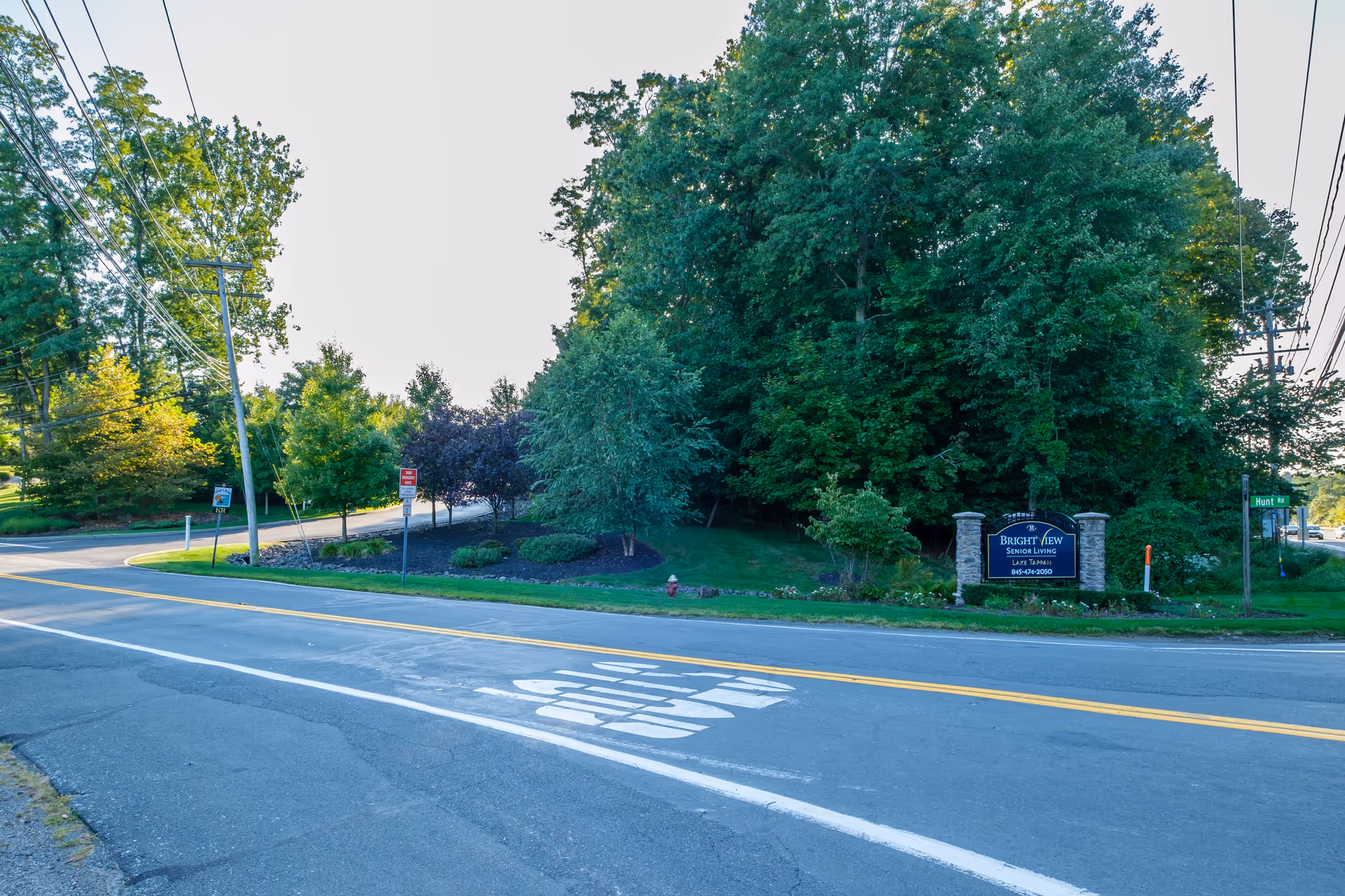 A street view showing the entrance to Brightview Lake Tappan Senior Living facility, with a sign on the right side surrounded by trees and greenery. The road has a painted 'STOP' marking, and there are utility poles and wires visible along the street.