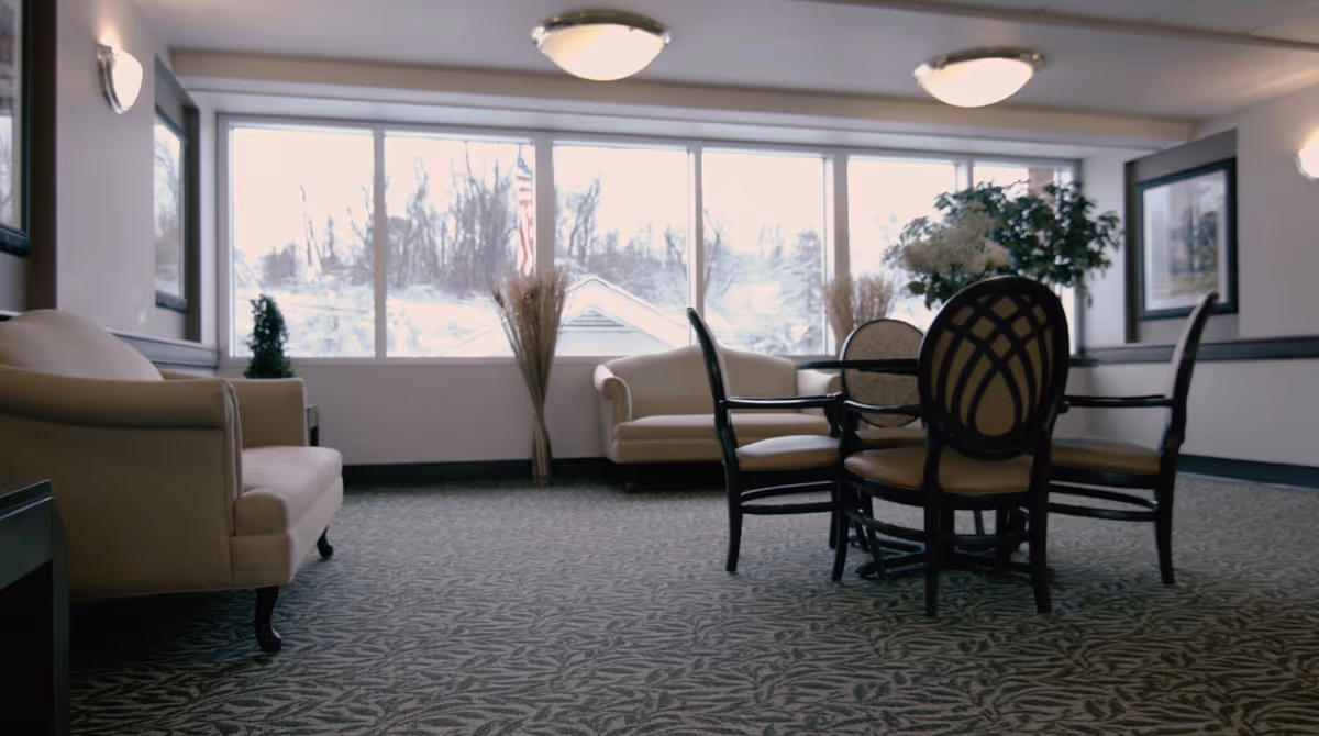 A bright common lounge with sofas, a round table and chairs in front of large windows.