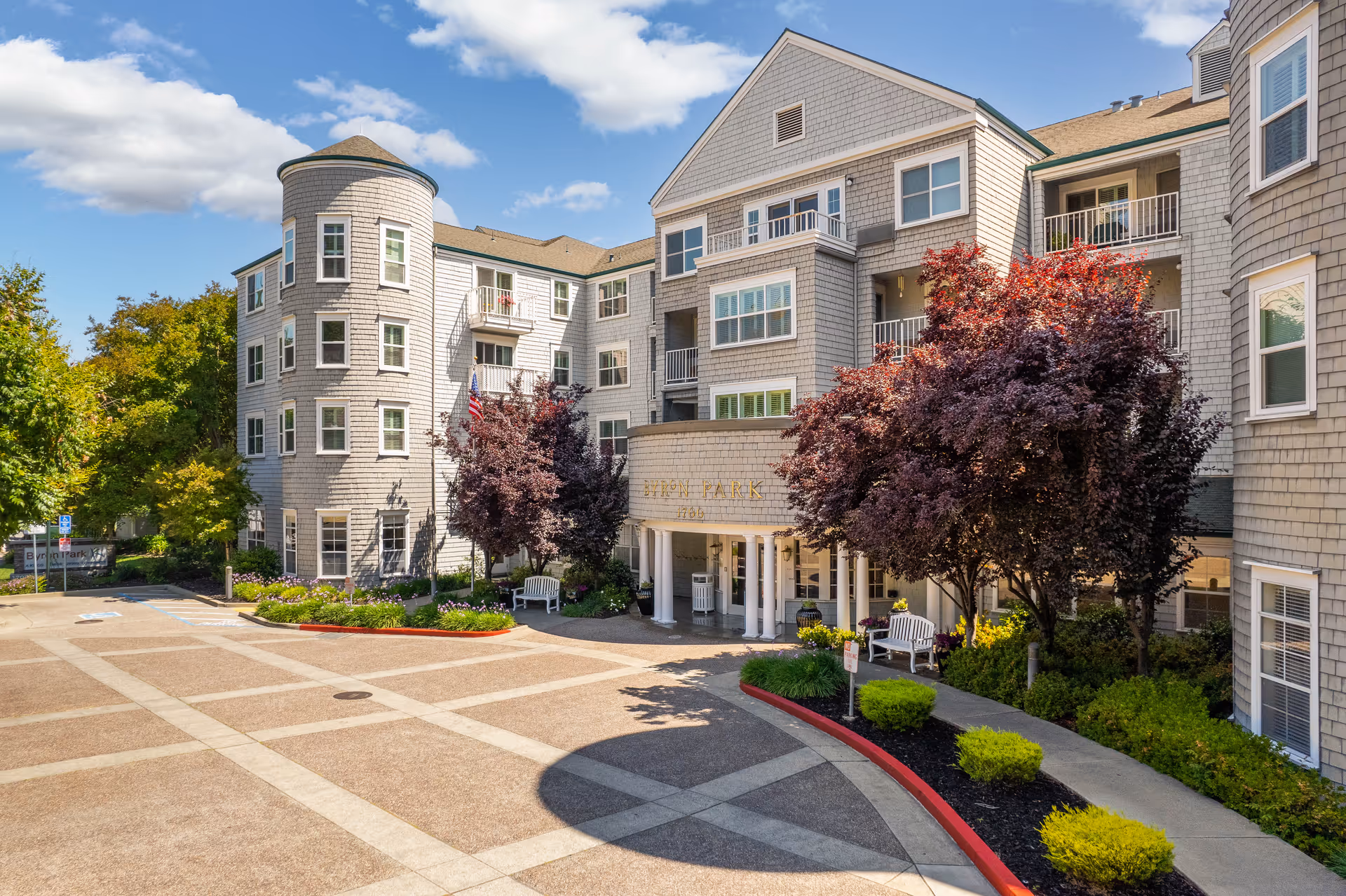 Exterior view of Byron Park senior living facility showing a multi-story building with gray siding, multiple windows, balconies, and a rounded tower section. The entrance is covered with white columns and surrounded by landscaped bushes and trees with red and green foliage. The sky is blue with some clouds.