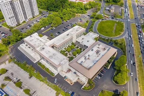 Aerial view of The Addison of Cherry Hill facility showing multiple connected buildings with flat roofs, surrounded by parking lots, trees, and nearby roads with vehicles.