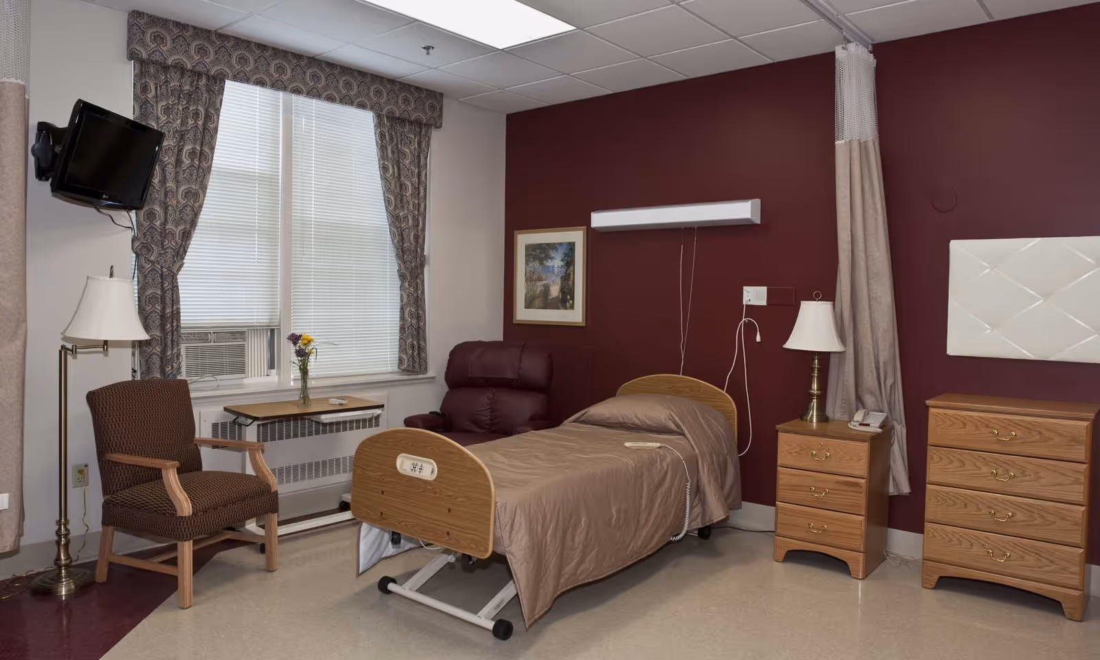 A furnished nursing facility bedroom with a single hospital-style bed, recliner, armchair, dressers, and a window with curtains.