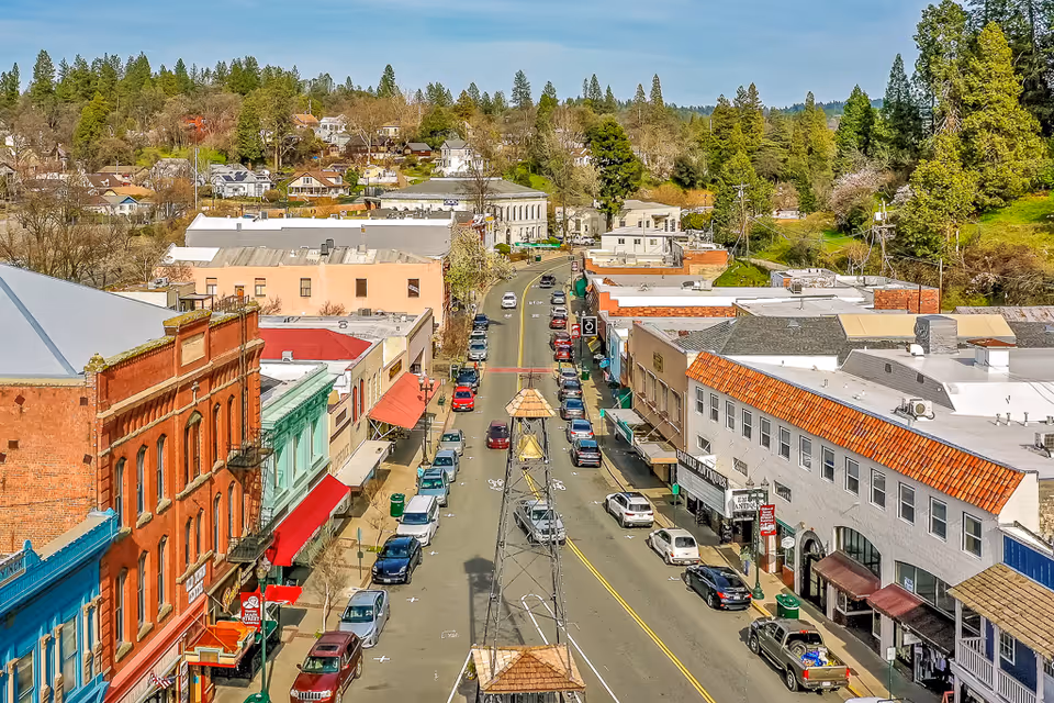Aerial view down a small-town main street lined with storefronts, parked cars, and trees in the background.