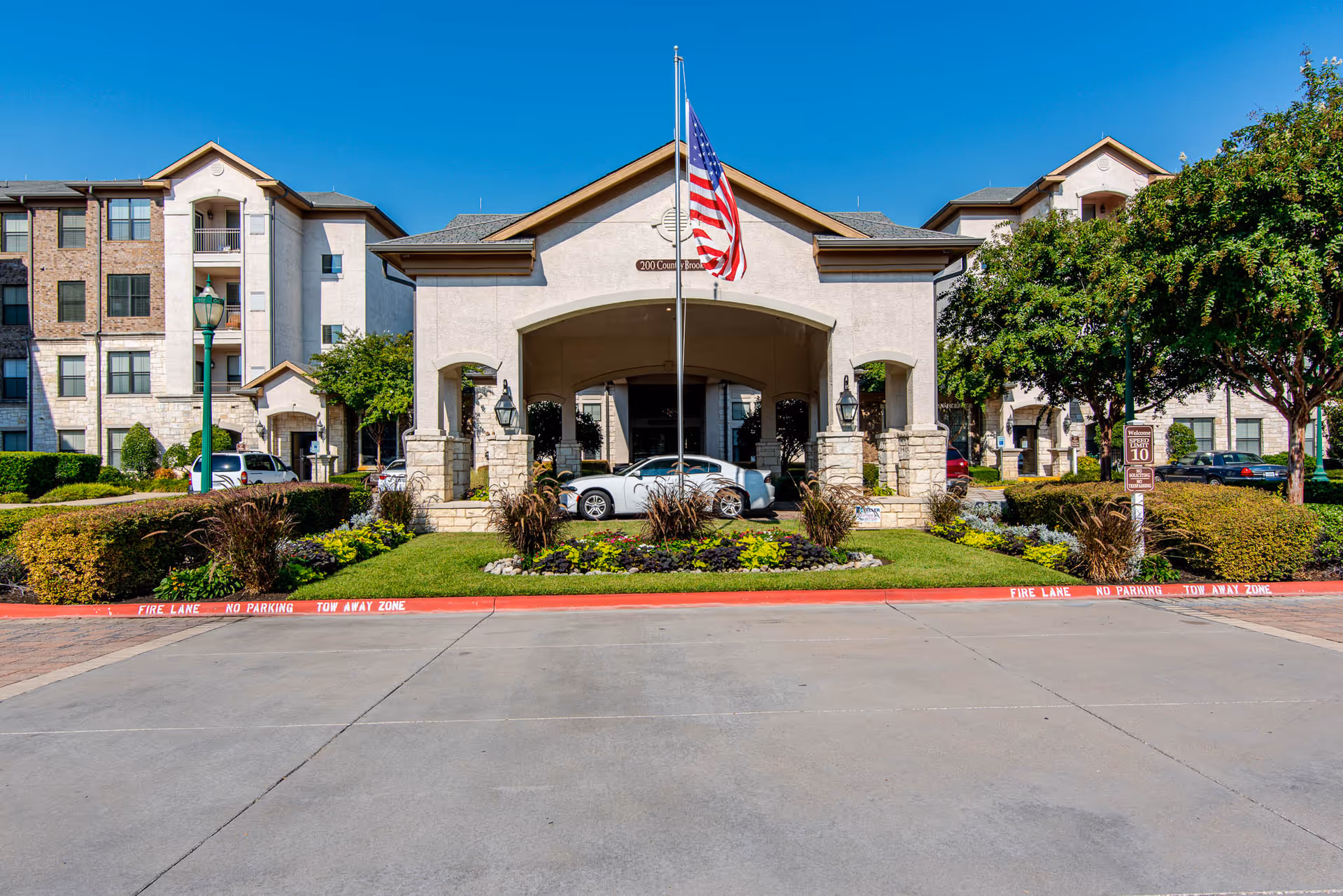 Front exterior view of Conservatory At Keller Town Center senior living facility with a covered entrance, an American flag on a flagpole, landscaped greenery, and parked cars.