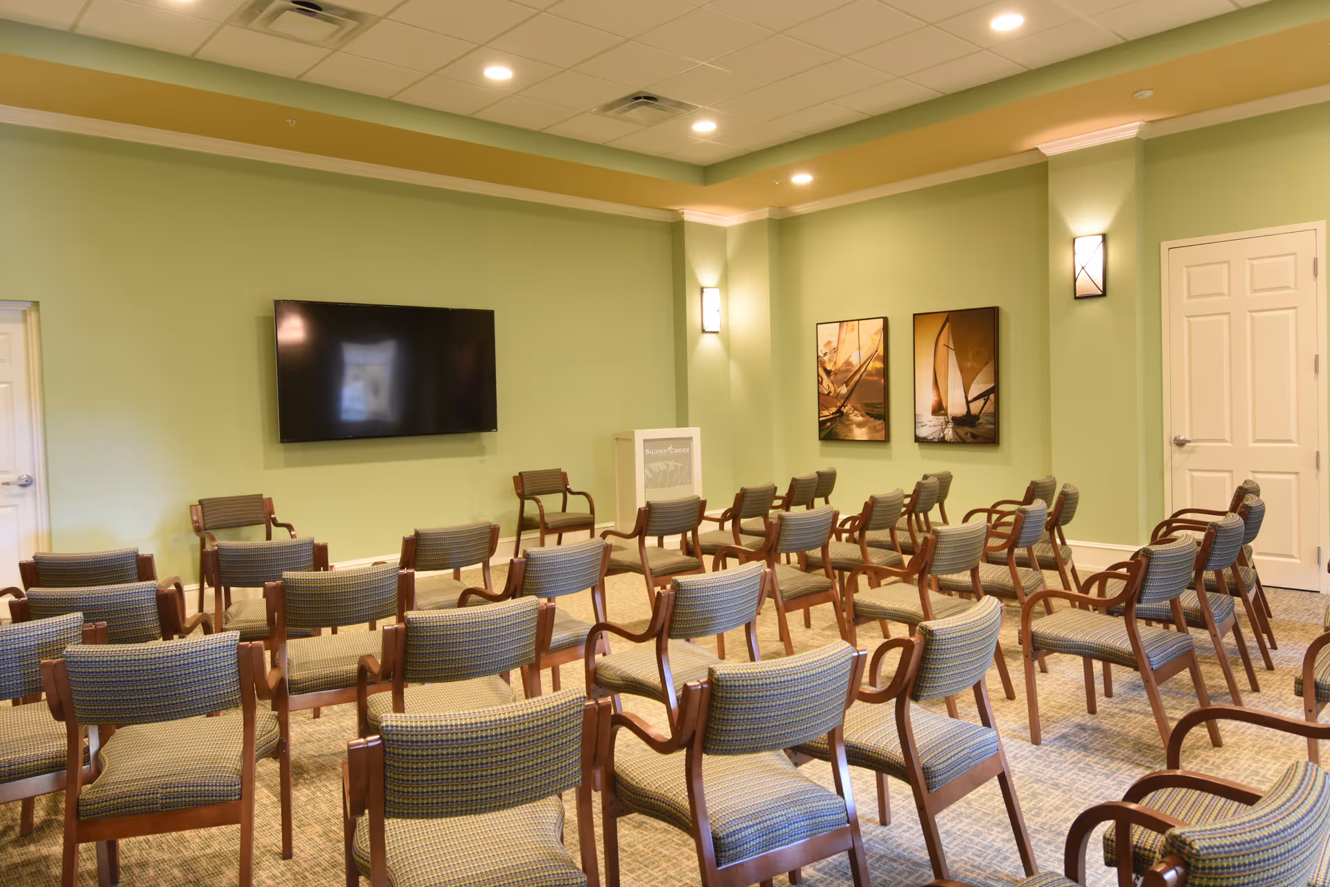 A green-walled common room with rows of chairs arranged facing a wall-mounted TV and a small podium.