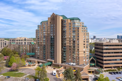 A tall multi-story residential building with a green roof surrounded by trees and other buildings under a partly cloudy sky.