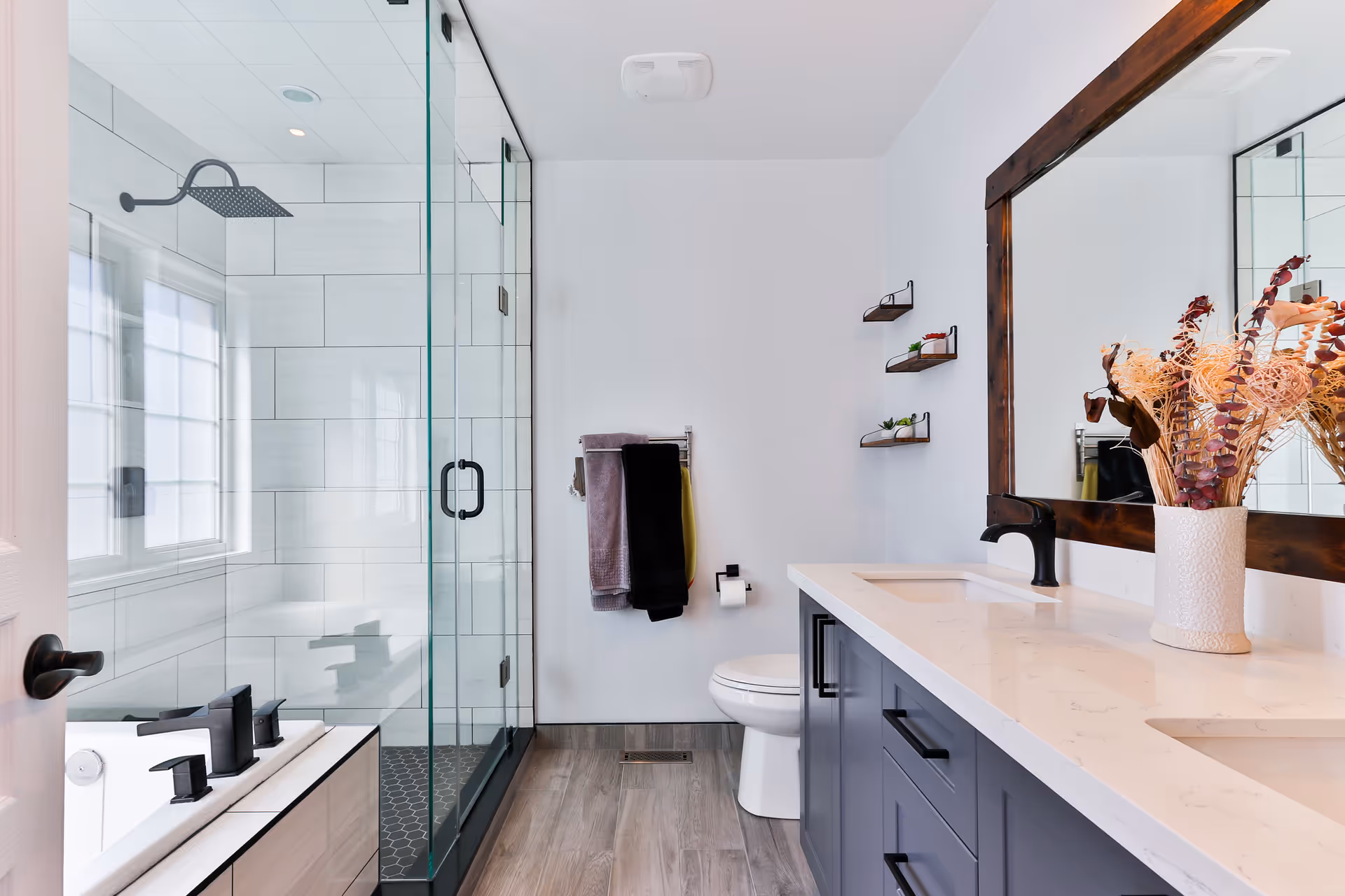 Modern bathroom with a glass-enclosed shower featuring a rainfall showerhead, a bathtub with black fixtures, a toilet, and a double sink vanity with a large wooden framed mirror and a vase with dried flowers. The walls are white, and the floor has wood-like tiles. Towels hang on a rack next to the toilet, and small shelves with plants are mounted on the wall.